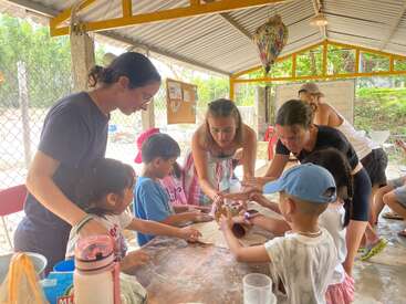 A group of adults and children are gathered around a table under a shelter, engaging in a fun hands-on activity, possibly making crafts or baking together.