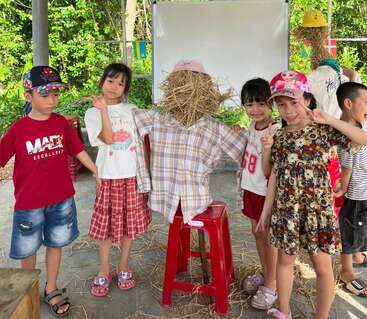 A group of smiling children stands around a homemade scarecrow made of straw and plaid shirt, posing together outdoors with greenery and a whiteboard behind them.