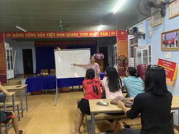 A teacher stands at a whiteboard, explaining a lesson to a small group of students seated in a brightly lit classroom with Vietnamese banners.