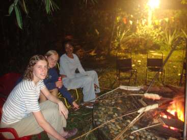 Three friends sit around a campfire at night, roasting food on sticks. They appear relaxed, enjoying the warmth, with festive lights and chairs in the background.