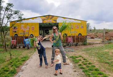 A woman and two children stand joyfully in front of a colorful barn labeled "Happy Farm," holding green plants on a cloudy day, surrounded by greenery.