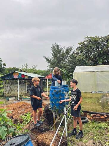 Three children are tending to a vertical blue barrel garden outdoors. They are laughing and working together in a lush, green garden with overcast skies.
