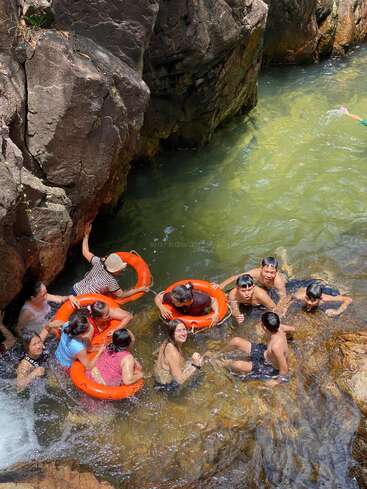 A group of people enjoy swimming and playing in a natural rocky pool, using orange floatation rings, surrounded by rocks and clear, greenish water. Laughter fills.