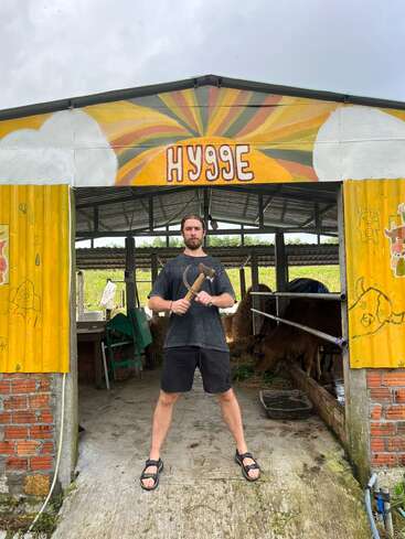 Un homme se tient, l'air assuré, devant une grange colorée sur laquelle est inscrit « HYGGE », un outil à la main. On aperçoit des animaux et du matériel à l'intérieur, sur fond de paysage rural.