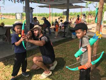 Un homme apprend à deux jeunes garçons à se servir d'un arc et de flèches factices en plein air. D'autres enfants et adultes se trouvent à l'ombre dans un parc.