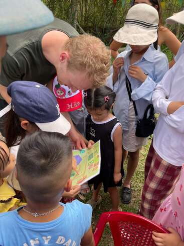 Un groupe d'enfants est rassemblé autour d'un adulte qui leur montre une carte ou un livret coloré en plein air ; ils participent ensemble à une activité à la fois éducative et ludique.