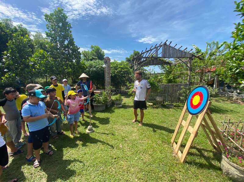 The image depicts a group of children and an adult standing in a grassy area, engaged in archery practice with bows and arrows, near a target on a wooden stand.