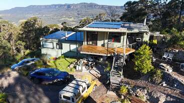 Une maison moderne équipée de panneaux solaires est située sur une colline entourée d'arbres, avec trois voitures garées, une terrasse en bois, des montagnes et un paysage pittoresque en arrière-plan.