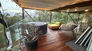 Cette terrasse extérieure confortable dispose d'un jacuzzi, de chaises longues confortables, d'une plante en pot, de guirlandes lumineuses et d'une magnifique vue sur la forêt, le tout entouré d'une balustrade en verre.