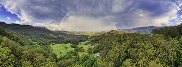 Une vallée verdoyante s'étend sous des forêts denses et des collines ondulantes. Des nuages spectaculaires remplissent le ciel, illuminés par la lumière du soleil, avec un arc-en-ciel pâle au-dessus.