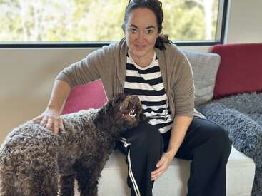 Une femme est assise sur un canapé moderne, souriant chaleureusement, vêtue d'une chemise rayée et d'un cardigan, tout en caressant affectueusement un joyeux chien brun aux poils bouclés à côté d'elle.