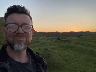 The image depicts a man with short hair and glasses, standing in front of a serene landscape featuring rolling hills and a sunset.