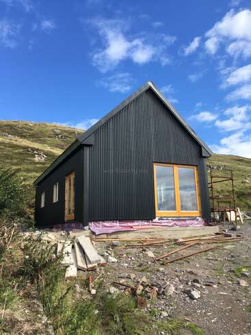 The image depicts a small, one-story house under construction, featuring a dark gray exterior, yellow-framed windows, and a sloping roof, set against a hillside backdrop.