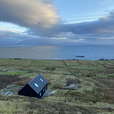 The image depicts a serene coastal landscape with a small black house, fields, and a body of water under a cloudy sky, evoking a sense of tranquility.