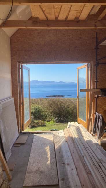 The image depicts a room under construction, featuring an open door with a scenic view of the ocean and mountains on a sunny day.