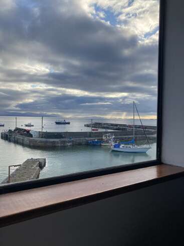 The image depicts a serene harbor scene, featuring a window frame in the foreground, with boats docked in the water and a cloudy sky above.
