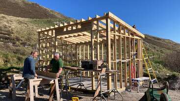 Dois homens trabalham na estrutura de uma casa de madeira sob um céu azul claro, cercado por colinas. Você pode ver uma escada amarela e várias ferramentas de construção nas proximidades.