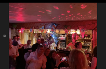 Uma cena de festa animada em um bar com iluminação quente, pessoas dançando, rindo, decorações, uma bola de discoteca, tema tropical e alguém capturando a diversão com um telefone.