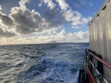 Um oceano agitado com ondas espumosas, uma ilha distante no horizonte, um céu nublado dramático, a luz do sol rompendo e uma grade de metal de barco em primeiro plano.