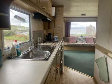 Esta imagem mostra uma cozinha de caravana bem arrumada com pia, fogão e material de limpeza, com vista para uma área de lounge aconchegante com janelas grandes e vistas panorâmicas do exterior.