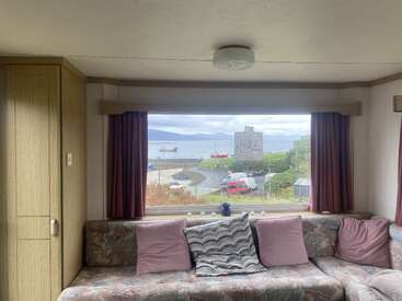 A foto mostra uma sala de estar aconchegante com sofá estampado, almofadas cor-de-rosa e uma grande janela com vista para o mar, barcos, uma torre e colinas.