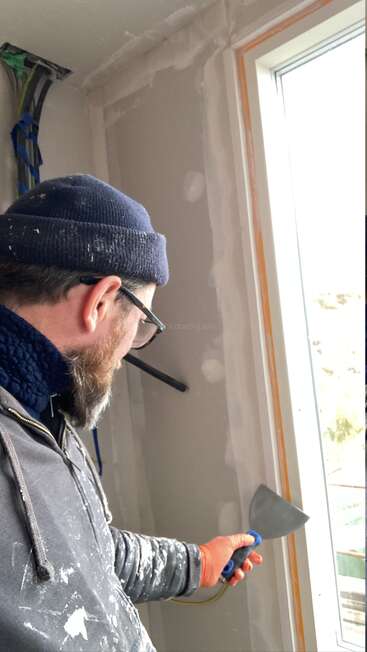 Um homem usando um gorro, óculos e um capuz manchado de tinta está trabalhando em um local fechado perto de uma janela, segurando uma espátula, possivelmente trabalhando com drywall.