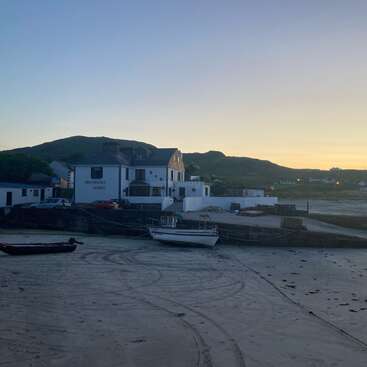 Uma cena costeira tranquila ao pôr do sol mostra barcos descansando em praias arenosas, um porto calmo, edifícios brancos, montanhas distantes e uma atmosfera tranquila e serena.