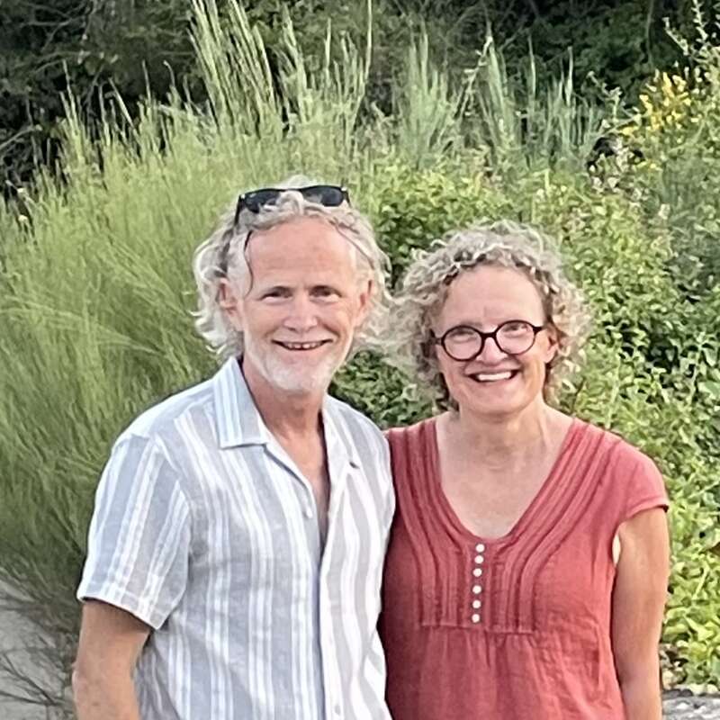 A smiling man and woman stand close together outdoors. Both have curly gray hair, and the woman wears glasses. Lush greenery fills the peaceful natural background.