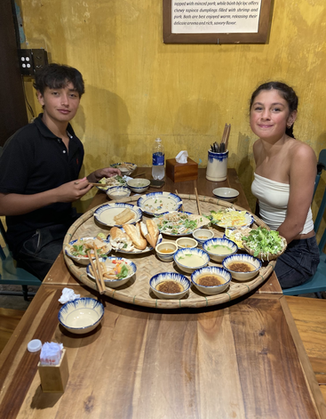 Zwei junge Leute sitzen an einem Holztisch, genießen ein reichhaltiges, farbenfrohes vietnamesisches Essen mit verschiedenen Gerichten, Dip-Saucen, Gemüse und Brot und lächeln glücklich ineinander.