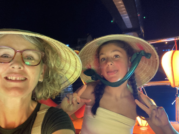 Zwei Menschen mit traditionellen Kegelhüten lächeln nachts für ein Selfie, während hinter ihnen bunte Laternen leuchten. Einer macht ein Peace-Zeichen und eine lustige Grimasse.