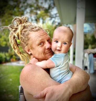 Un homme aux dreadlocks blondes serre tendrement dans ses bras un bébé aux yeux bleus brillants. Ils sont à l'extérieur et regardent tous deux l'appareil photo avec amour, partageant un moment sincère.
