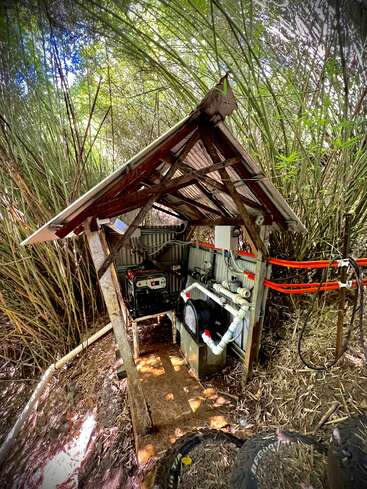 Dans une forêt de bambous luxuriante, un petit hangar métallique abrite un générateur et divers tuyaux. Des câbles orange vif et de gros pneus sont visibles sur le sol à proximité.