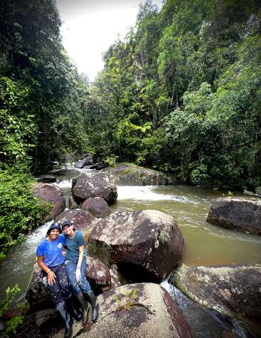 Deux personnes sont assises sur de gros rochers au bord d'une rivière qui coule, entourée d'une forêt tropicale verdoyante. L'atmosphère est paisible, sauvage et aventureuse, évoquant l'exploration et l'amitié.