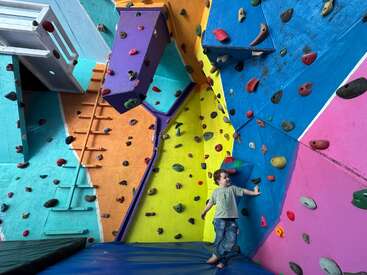 Un jeune enfant se tient pieds nus sur un tapis rembourré dans une salle d'escalade colorée, entourée de murs d'escalade multicolores et de nombreuses poignées.