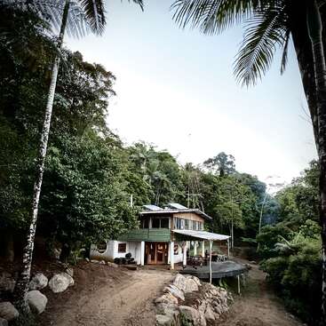 Una casa rústica de dos plantas está enclavada en una selva frondosa y densa. La rodean palmeras y rocas, y un camino de tierra conduce a la entrada. Un refugio tranquilo.