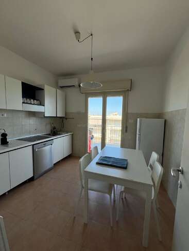 Dieses Bild zeigt eine moderne Küche mit weißen Schränken, Geschirrspüler, Esstisch und vier Stühlen, Kühlschrank und einer großen Glastür, die auf einen Balkon führt.