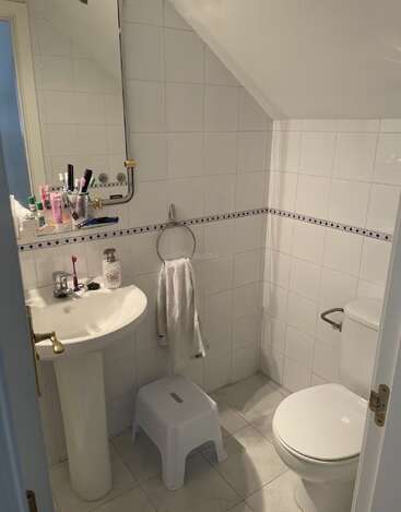 Dies ist ein kleines, weiß gefliestes Badezimmer mit einem Waschbecken, einem Spiegel, einer Toilette, einem Handtuchring, einem Tritthocker, Toilettenartikeln und einer schrägen Decke über der Toilette.