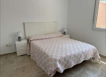 Ein einfaches, aufgeräumtes Schlafzimmer mit einem Doppelbett, einer gemusterten rosa-weißen Bettdecke, zwei Nachttischen, Lampen, einem weißen Kopfteil, einem gefliesten Boden und einem Fenster. Friedliche Atmosphäre.