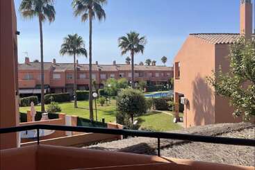 Dieses Bild zeigt einen sonnigen Innenhof mit Palmen, einem Swimmingpool, grünem Gras und terrakottafarbenen Wohnhäusern, betrachtet von einem Balkon mit Geländer.