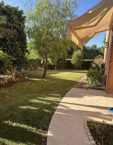 Ein sonniger Garten im Hinterhof mit grünem Gras, verschiedenen Bäumen und Sträuchern. Neben dem Haus befindet sich eine geflieste Terrasse, die von einer großen beigen Markise beschattet wird. Friedliche Szene.