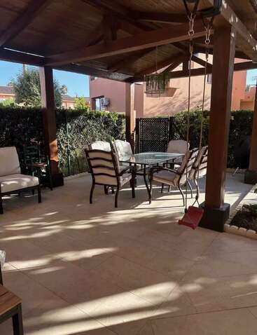 Dieses Bild zeigt eine gemütliche Terrasse mit gepolsterten Stühlen um einen Glastisch, der von einer hölzernen Pergola beschattet wird. Im Vordergrund hängt eine rote Schaukel.