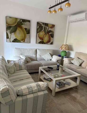 In diesem gemütlichen Wohnzimmer gibt es drei Sofas, Wandkunst mit Obstmotiven, einen gläsernen Couchtisch mit Zeitschriften, gestreifte Kissen, eine grüne Lampe, geflieste Böden und eine Deckenbeleuchtung.