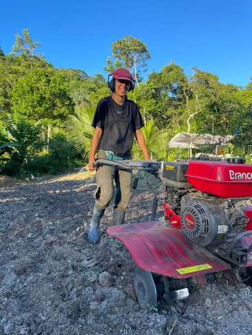 Une personne vêtue d'une tenue de travail et portant des protections auditives utilise un motoculteur rouge dans un champ fraîchement labouré, au milieu d'arbres verdoyants et sous un ciel bleu.