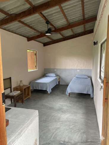 Cette photo montre une chambre simple et rustique équipée de deux lits simples, d'une parure de lit à carreaux bleus, d'un plafond en bois, d'un sol en béton, de meubles en bois, d'une fenêtre et d'un ventilateur de plafond.
