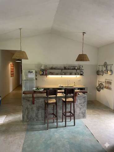 Cette cuisine minimaliste se caractérise par des plans de travail en béton, des tabourets de bar en bois, des suspensions en osier, des étagères ouvertes où sont disposés de la vaisselle, des casseroles suspendues, des ustensiles et un réfrigérateur, le tout dans des tons neutres.