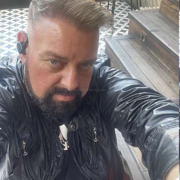 La imagen muestra a un hombre con pelo corto y barba, vestido con una chaqueta negra, tomándose un selfie en una terraza de madera con suelo de baldosas estampadas.