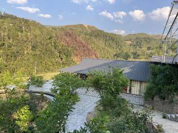 The image depicts a serene mountainous landscape featuring a building with a tiled roof, surrounded by lush greenery and a stone pathway, set against a blue sky with white clouds.