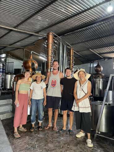 The image depicts five individuals standing in a distillery, posing for a photo, with copper and stainless steel equipment visible in the background.