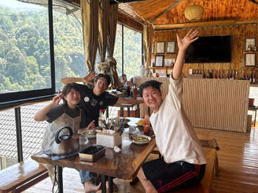 Three friends sit around a wooden table in a cozy, sunlit restaurant with large windows, smiling and posing playfully, enjoying food and a scenic mountain view.