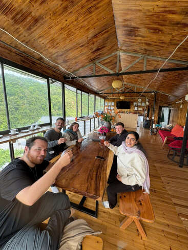 Five friends sit around a rustic wooden table in a cozy, wooden cabin with big windows, enjoying drinks and laughter, surrounded by lush green hills outside.